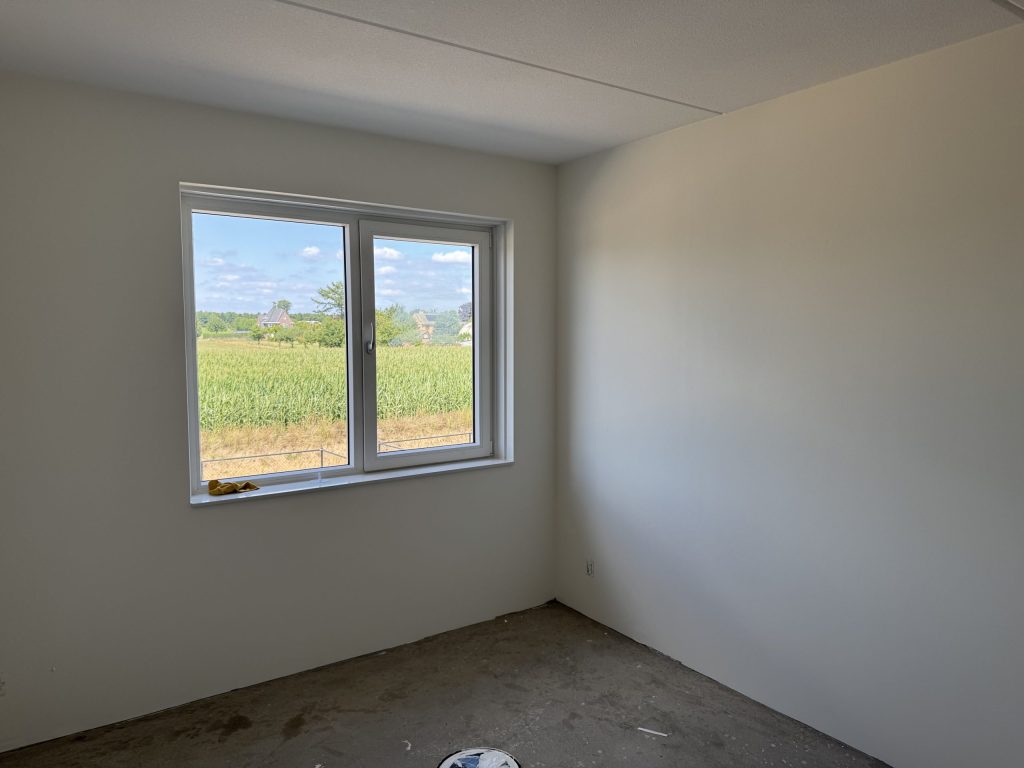 Lege slaapkamer in een nieuwbouwwoning met lichte wanden en een raam met uitzicht op een maisveld
